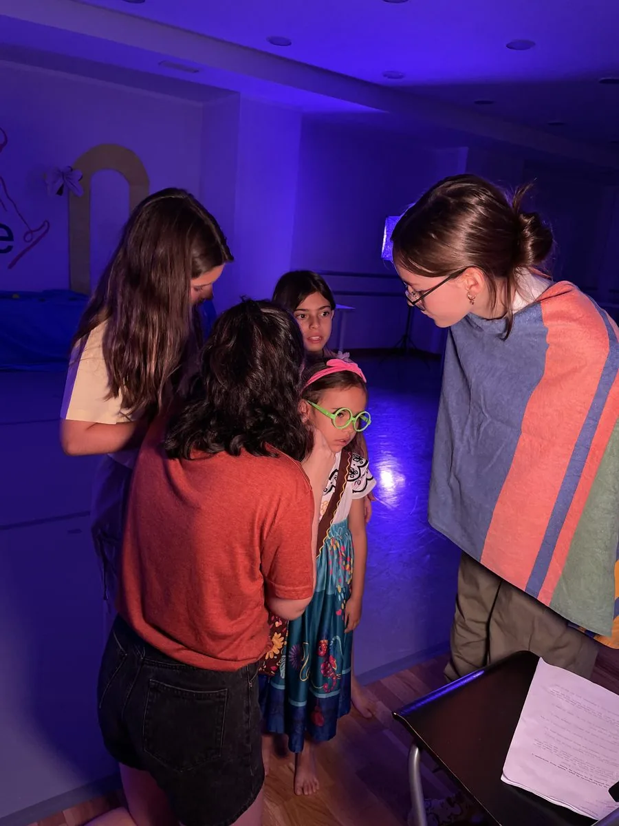 Monitoras y adolescentes rodean a una niña con gafas de colores mientras preparan el vestuario del musical