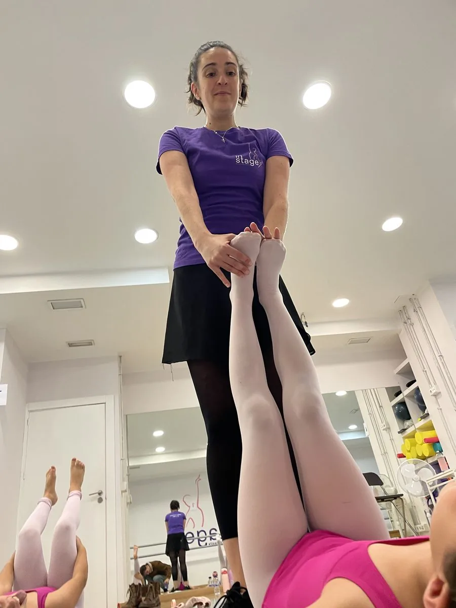 Profesora de In Stage con camiseta morada sujetando las piernas estiradas de una alumna tumbada boca arriba, en un trabajo de flexibilidad en la sala