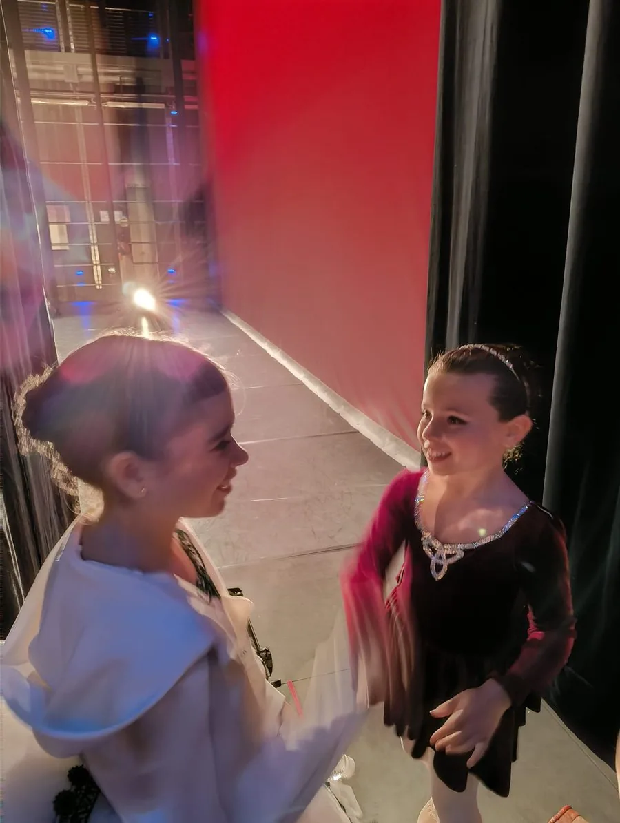 Dos bailarinas de In Stage entre bastidores, una con capa blanca y otra de negro, riéndose bajo la luz roja justo antes de salir a escena