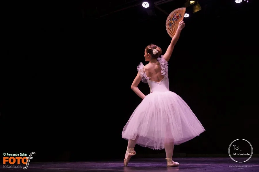 Bailarina de espaldas con tutú rosa, en puntas y con un abanico en alto, sola en el escenario