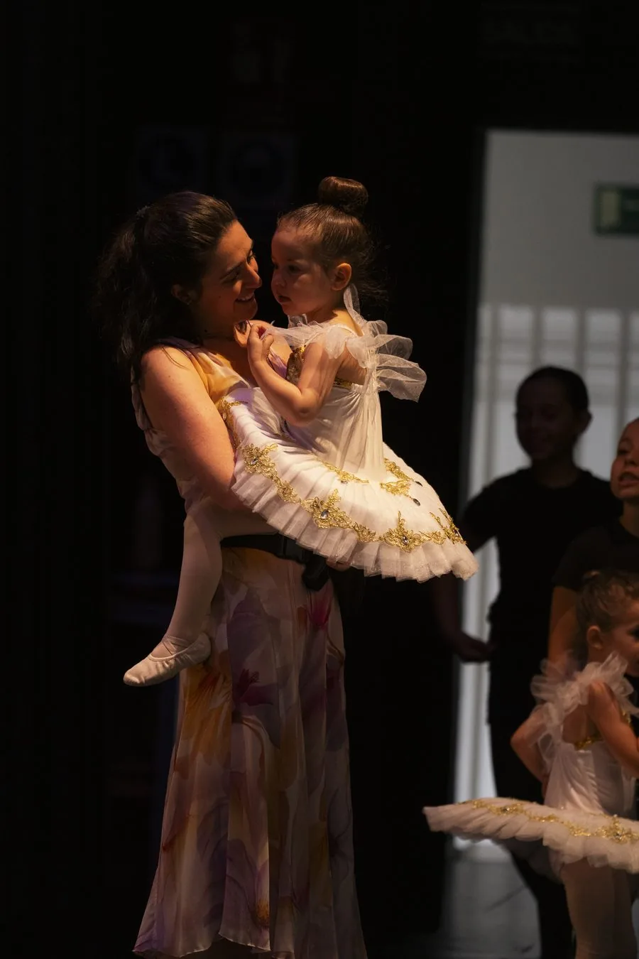 Profesora de In Stage con una niña pequeña en brazos, vestida con tutú blanco, mirándose las dos entre bastidores antes de salir a escena