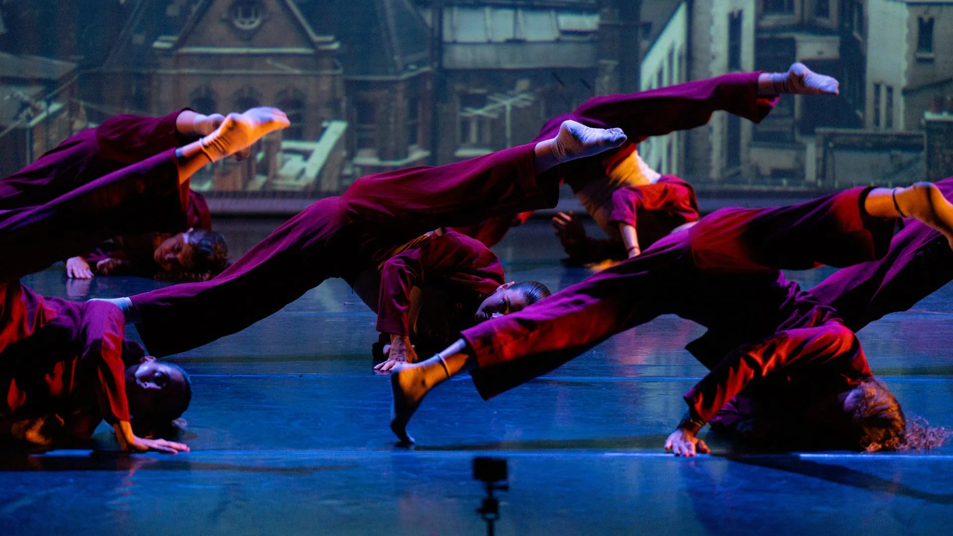 Grupo de bailarinas de contemporáneo con monos granates en una pose baja de conjunto, con luz azul sobre el escenario